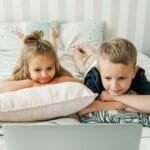 Dwoje dzieci leży na łóżku i ogląda laptop w przytulnym pokoju