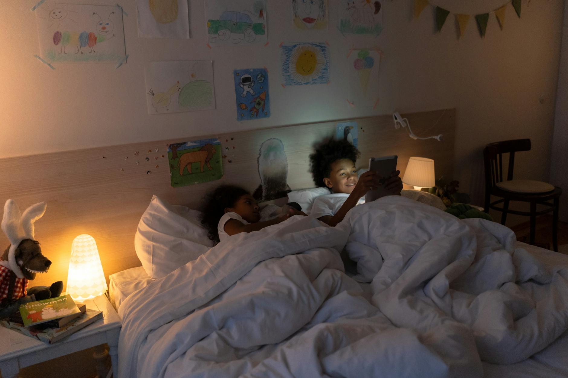 Dwoje dzieci ogląda tablet w przytulnej, ciepło oświetlonej sypialni